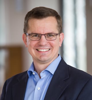 Professional headshot of a man wearing glasses, blue shirt, and dark blazer, smiling at camera against blurred office background