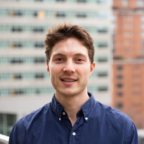 Smiling man wearing a dark blue button-up shirt, photographed outdoors with urban apartment buildings in the background