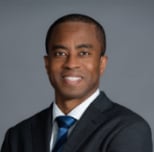 Professional headshot of a man in a dark blazer and blue tie against a neutral gray background
