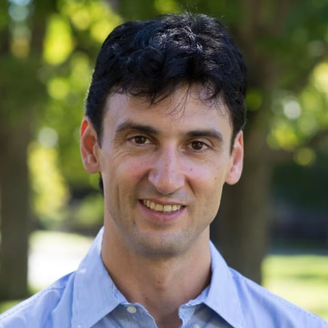 Itai Yanai smiling in a light blue button-up shirt outdoors with blurred green trees in the background