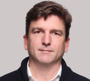 Headshot of a middle-aged man with brown hair wearing a black jacket over a white shirt against a gray background