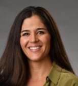 Professional headshot of a woman with long dark hair smiling at the camera, wearing a olive green top