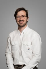 Man with brown hair and glasses wearing a white button-up shirt, smiling at the camera against a gray background.
