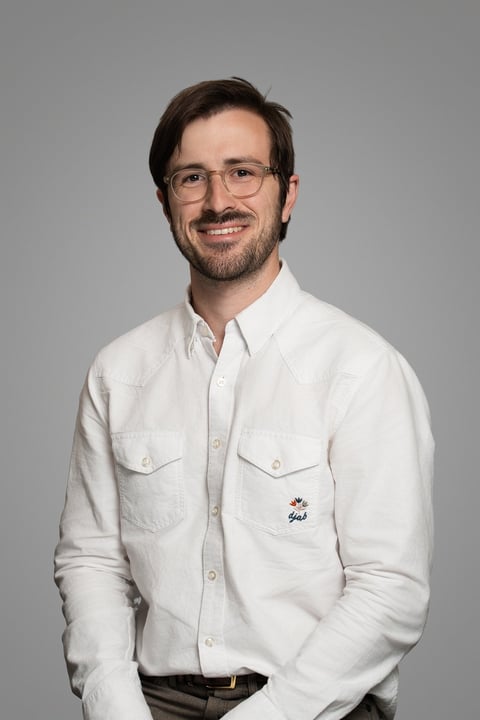 Man with brown hair and glasses wearing a white button-up shirt, smiling at the camera against a gray background.