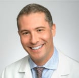 Professional headshot of a smiling man in a white coat and light blue shirt against a neutral gray background