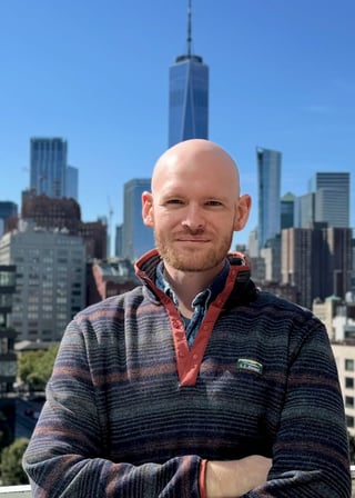 Bald man in striped pullover smiling at camera with NYC skyline and One World Trade Center in background