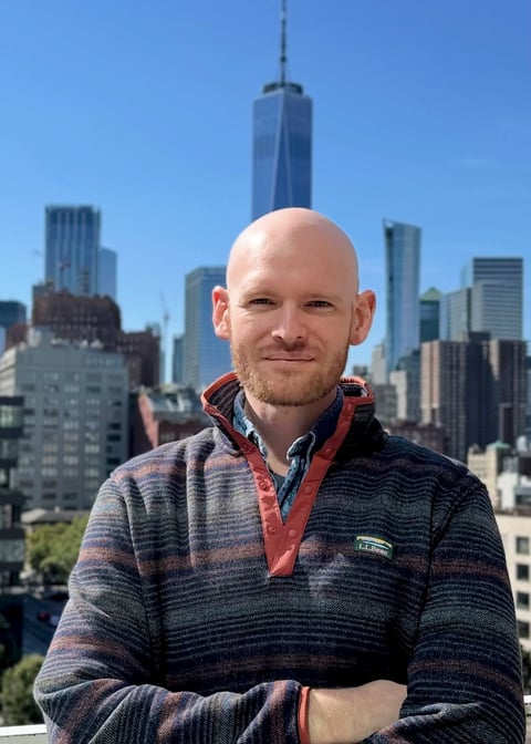 Bald man in striped pullover smiling at camera with NYC skyline and One World Trade Center in background
