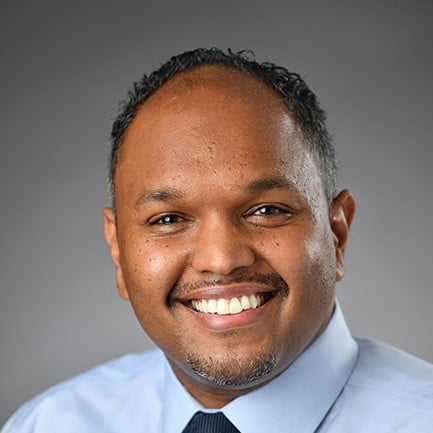 Professional headshot of a man wearing a light blue shirt and dark tie, smiling against a gray background