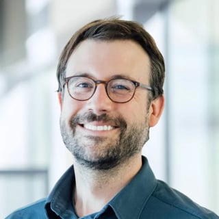 Portrait of a smiling man with glasses and a beard wearing a dark blue shirt against a blurred office background