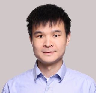 Portrait of a man with short black hair wearing a light blue button-up shirt against a neutral gray background